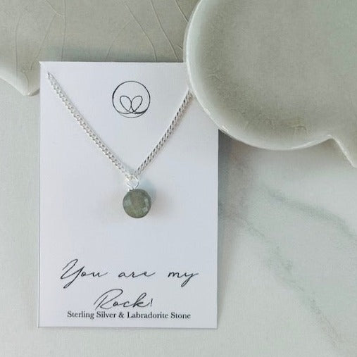 Sterling silver necklace with a round Labradorite stone, symbolizing strength and stability. Displayed on a 'You Are My Rock' background. Includes an 18” curb chain.